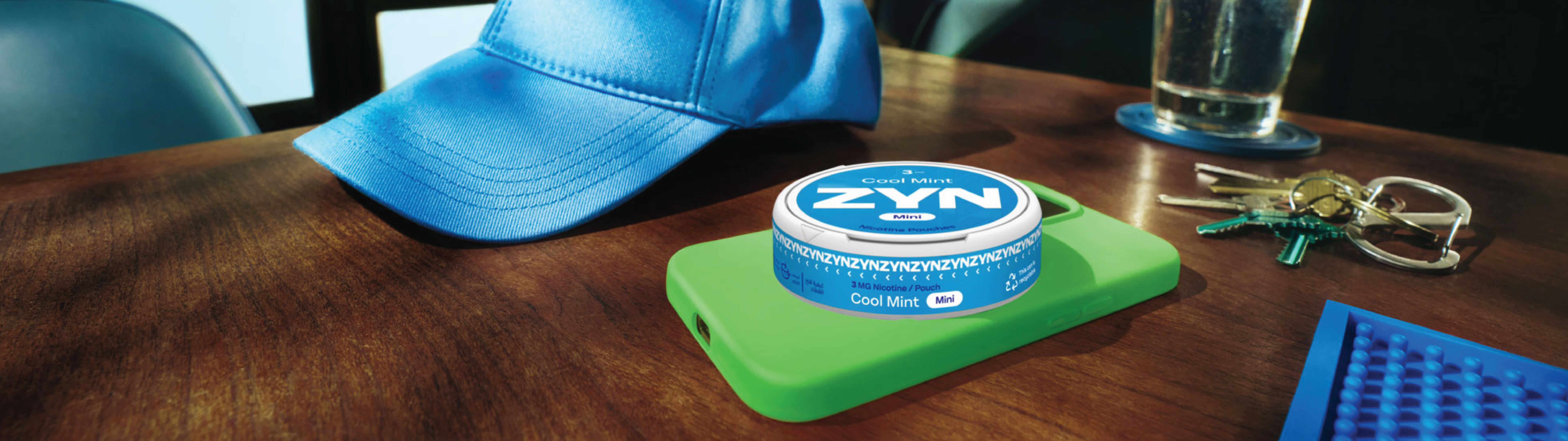 A ZYN container placed on top of a phone with a green case next to a blue cap, keys and a drink on a brown table